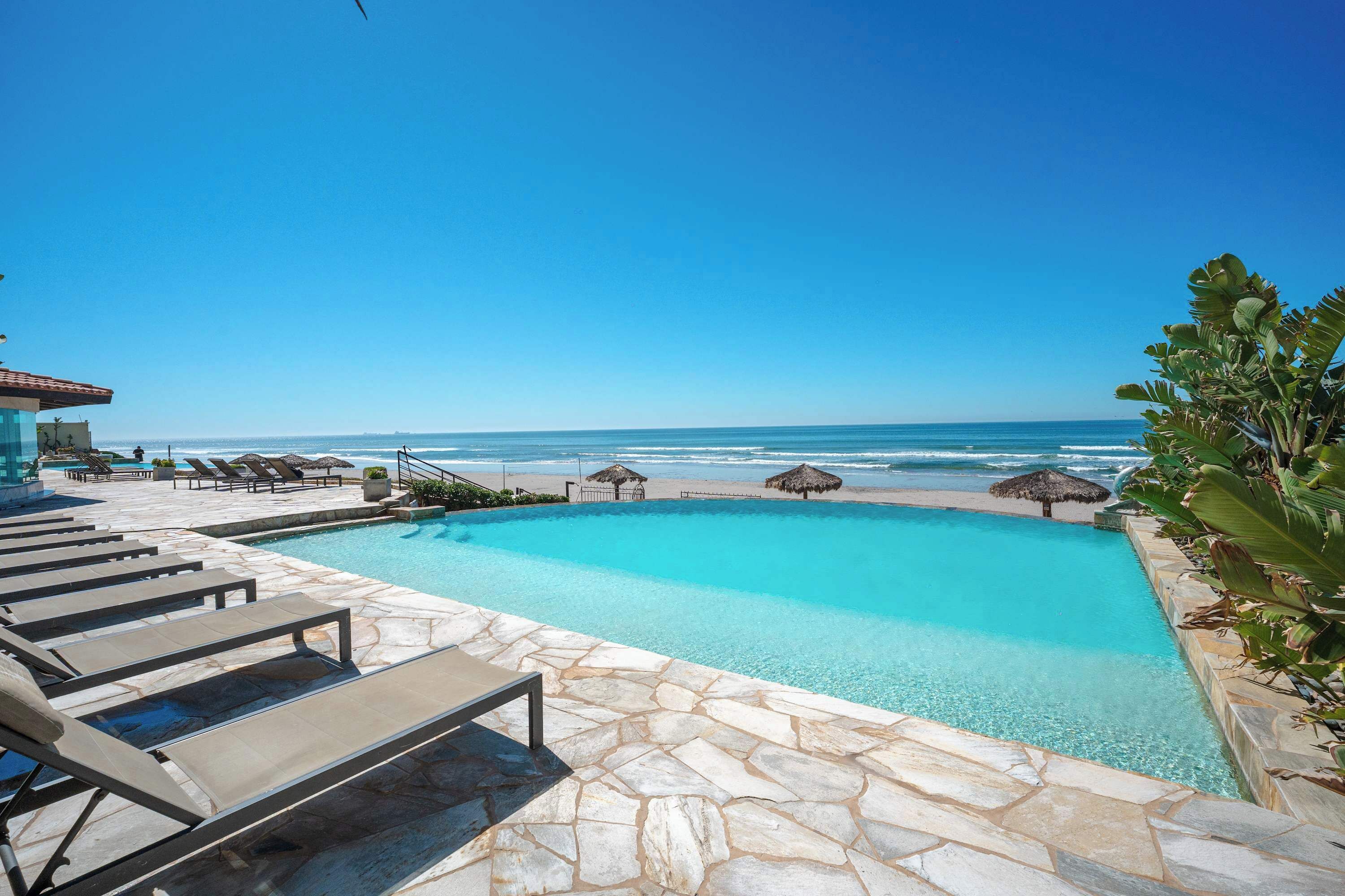A pool with a view of the beach, lounge chairs, and a building with a tiled roof.