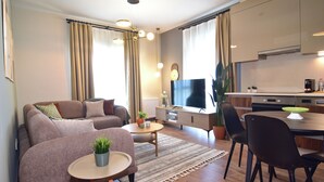 Ein modernes Wohnzimmer mit Sofa, Couchtisch und Fernseher. Es gibt einen Essbereich mit Stühlen und Tisch. Der Raum verfügt über eine Küche mit Schränken und Geräten.