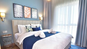 Ein ordentlich bezogenes Bett mit weißer und blauer Bettwäsche, einer blauen Tagesdecke und zwei aufgerollten Handtüchern. Zwei Wandlampen, ein runder Beistelltisch und ein großes Fenster mit durchsichtigen Vorhängen sind ebenfalls vorhanden.