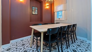 Un comedor con mesa de madera y sillas negras, un aplique de pared y una ventana con persianas.