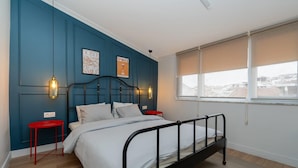 Un dormitorio con estructura de cama metálica negra, ropa de cama blanca, un taburete rojo y dos lámparas fijadas a la pared.