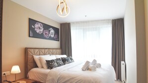 A neatly made bed with pillows and towels, a bedside lamp, and a framed floral artwork on the wall.