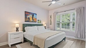 A bedroom with a bed, nightstands, a ceiling fan, a large window with curtains, and artwork on the wall. - Oh Gosh! So Posh in Myrtlewood Villas (Myrtle Beach)