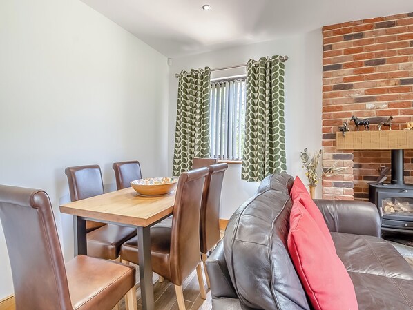 Un coin repas avec une table en bois et des chaises en cuir marron, un salon avec un canapé en cuir et une cheminée, et des rideaux à motifs verts.