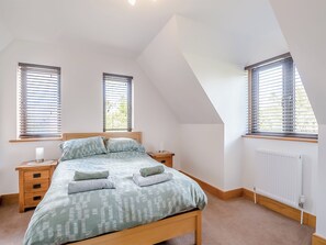 A bedroom with a sloped ceiling, two bedside tables with lamps, a bed with patterned bedding, and windows with blinds. - 3 bedroom accommodation in Lowestoft (Lowestoft)