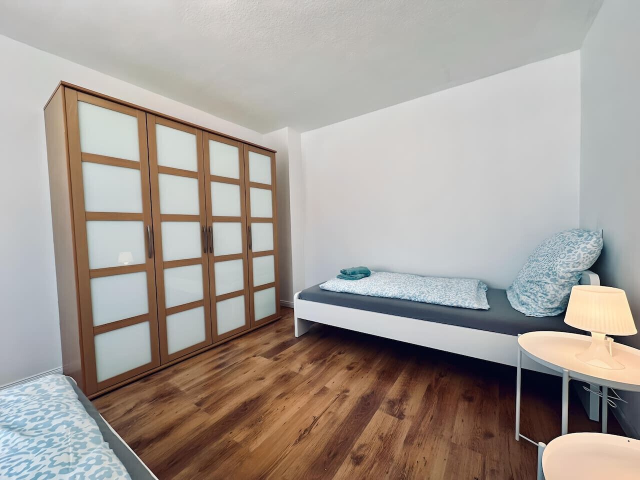 Een slaapkamer met een bed, nachtkastje, lamp, kledingkast en een bank met kussens.