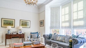 Een woonkamer met een bank, een bijzettafel met boeken, een gestreept fauteuil en een kroonluchter.