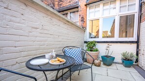 Een klein terras buiten met een ronde metalen tafel, twee stoelen, een wit bord met eten, een kopje en twee plantenpotten.