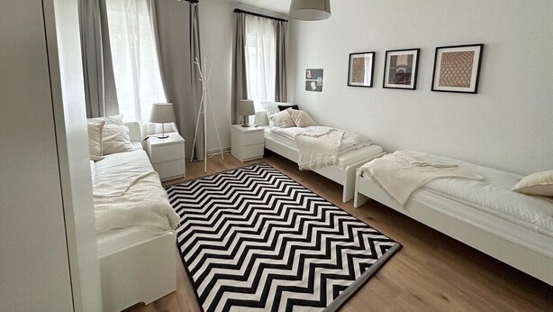 Une chambre moderne avec deux lits simples, un grand tapis à motifs chevrons noir et blanc, une lampe sur pied et des tableaux encadrés accrochés au mur.