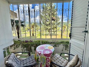 Een balkon met uitzicht op een tuin en bomen, een ronde tafel met bloemen tafelkleed, en twee comfortabele stoelen.