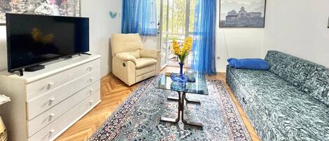 Una sala de estar con televisor, sofá, sillón y una mesa de centro de vidrio con flores.