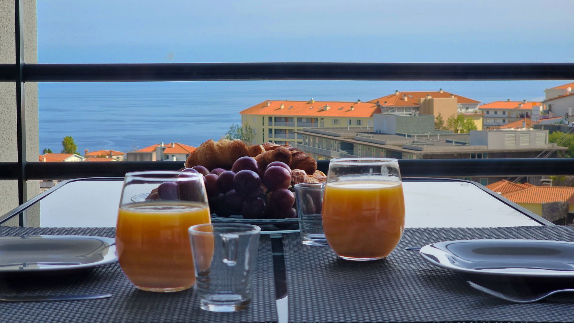 Une table dressée avec des aliments pour le petit-déjeuner et des verres de jus d’orange, donnant sur une ville côtière.