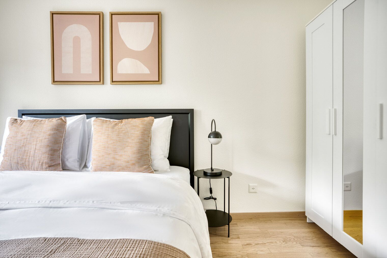 A bedroom with a bed, bedside table, lamp, wardrobe, and two framed artworks on the wall.