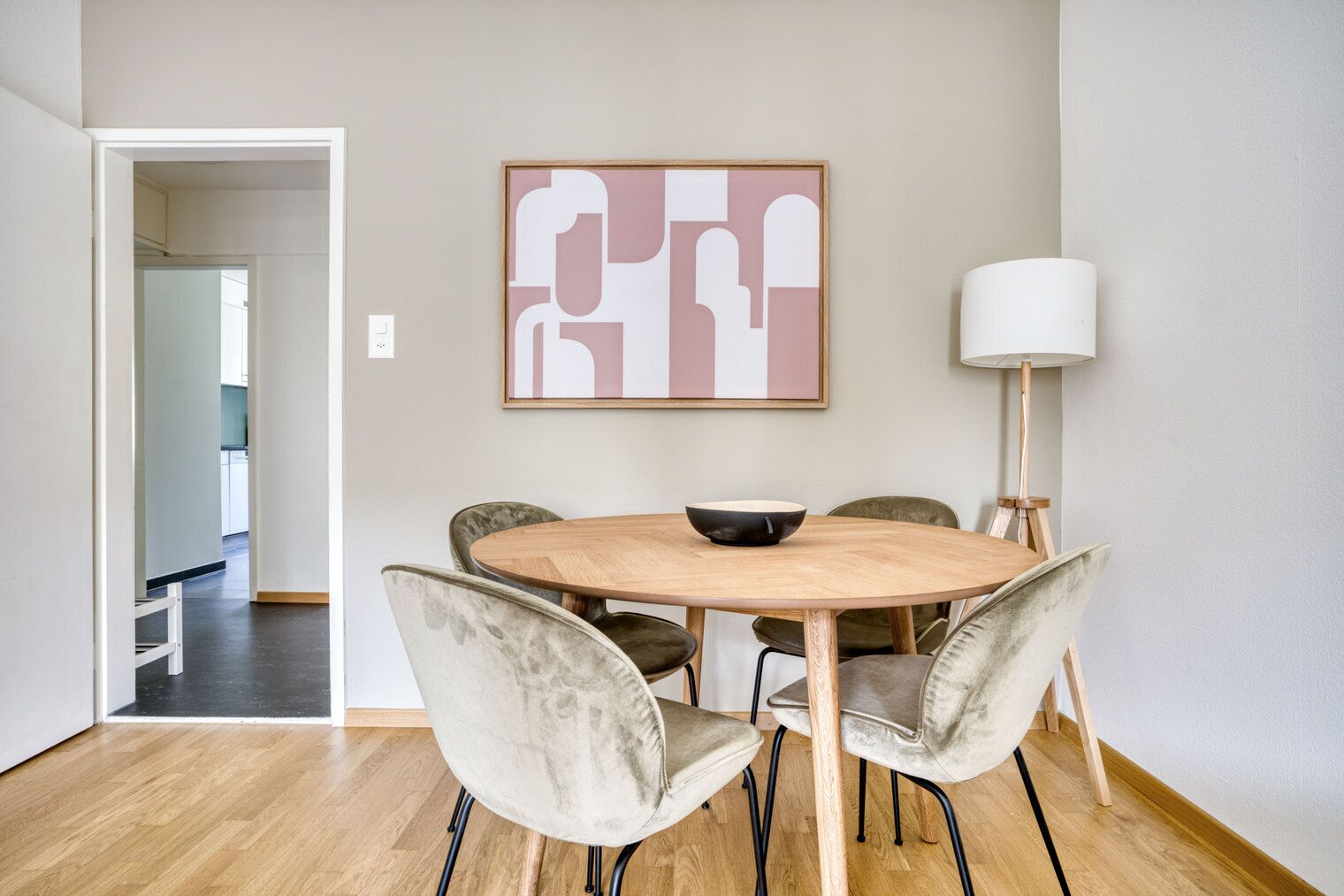 Un coin repas comprenant une table ronde en bois, quatre chaises, un saladier, un tableau abstrait accroché au mur et une lampe sur pied.