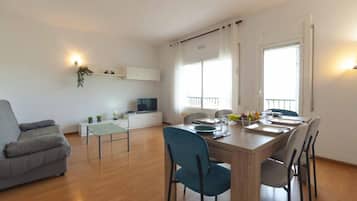 Una sala moderna con un sofá gris, una mesa de comedor con sillas, un televisor y un balcón con barandilla.