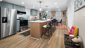 A modern kitchen with stainless steel appliances, a central island with bar stools, pendant lighting, and a dining area with a patterned rug.
