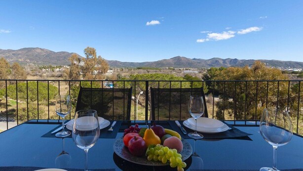 Taula per a dues persones amb un plat de fruita i gots de vi, amb vista a un paisatge de muntanyes.