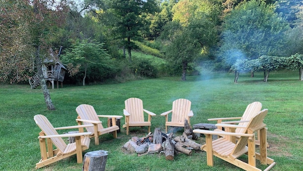A group of Adirondack chairs arranged around a fire pit in a grassy outdoor area with trees in the background.