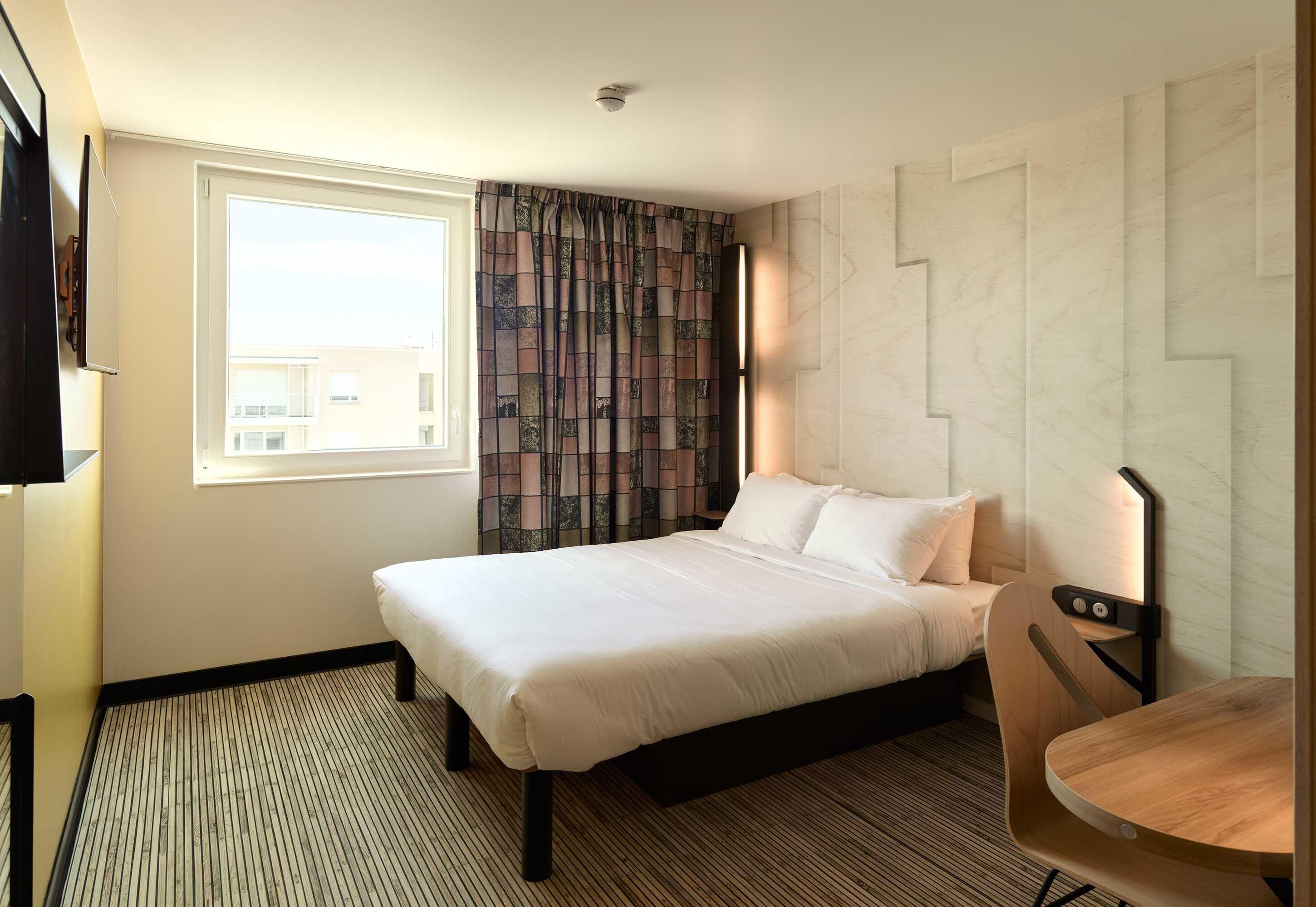A hotel room with a bed, a TV, a window with curtains, a wooden table, and a patterned wall.