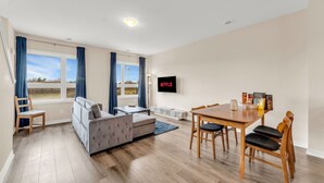 A modern living room with a sofa, a dining table with chairs, a TV mounted on the wall, and large windows with curtains. - Stylish Niagara Falls Getaway with Rooftop City Views (Niagara Falls)