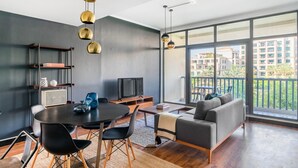 A modern living room with a round dining table, black chairs, a grey sofa, and a TV. There are shelves with decorative items and a large window offering a view of buildings and greenery. - Blueground | The Views, pool, nr Canal (Dubai)