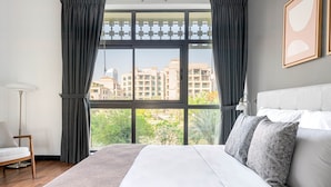 A modern bedroom with a large bed, grey curtains, a view of buildings and trees, and a floor lamp. - Blueground | The Views, pool, nr Canal (Dubai)