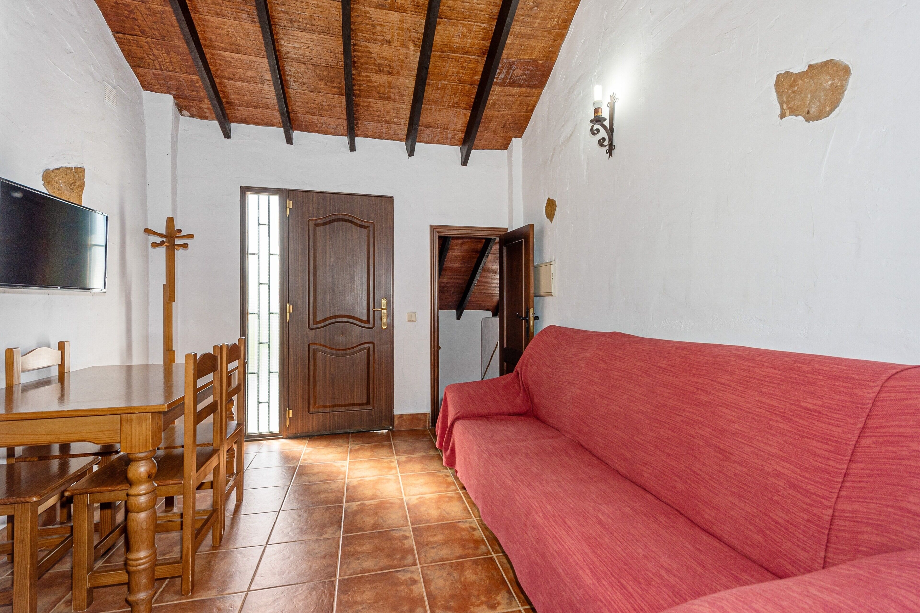 Ein Zimmer mit einem Holzesstisch, Stühlen, einem roten Sofa, einer Holztür und einem an der Wand befestigten Fernseher.
