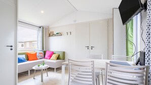 Una habitación moderna con un sofá blanco, una mesita, una mesa de comedor con sillas, un televisor montado en la pared y una ventana con vistas.