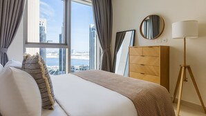 Habitación de hotel moderna con una cama grande, un aparador de madera, una lámpara de pie y vistas a un paisaje urbano a través de amplios ventanales.