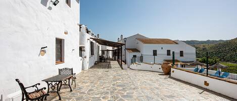 A stone-paved courtyard with outdoor furniture, white buildings, and a view of hills.