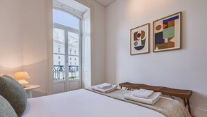 Une chambre d’hôtel avec un grand lit, une petite table avec une lampe, une fenêtre donnant sur des immeubles et deux tableaux abstraits accrochés au mur.