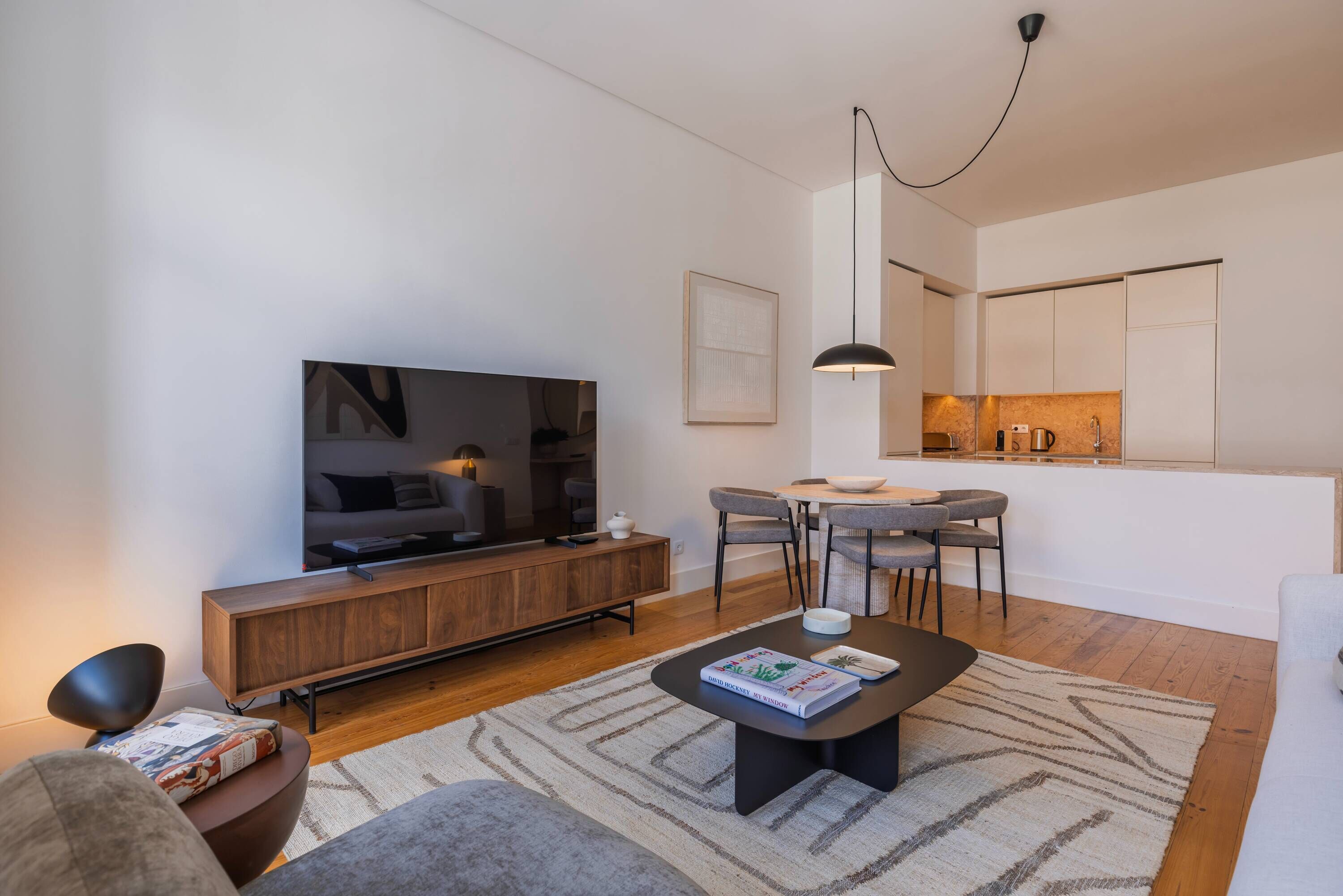 A modern living room with a flat-screen TV, wooden console, and a dining area with a kitchen in the background.