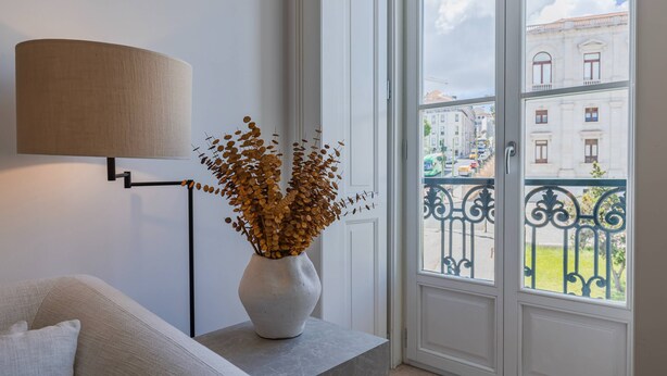 Une pièce comprenant un canapé, une lampe, un vase contenant des feuilles séchées et une fenêtre donnant sur des immeubles et une rue.