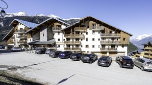A multi-story hotel with balconies, a parking lot, and a mountainous backdrop. - Nice apt near the slopes for 6 pers (Modane)