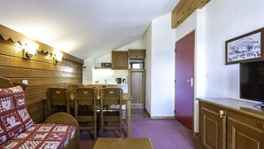 A compact living space with a wooden dining area, a sofa with patterned cushions, a kitchen with basic appliances, and a television on the right. - Nice apt near the slopes for 6 pers (Modane)