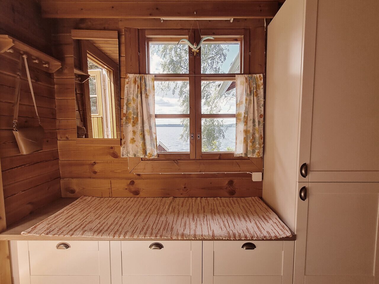 A cabin interior with a wooden bench, a window with curtains, and a white cabinet.