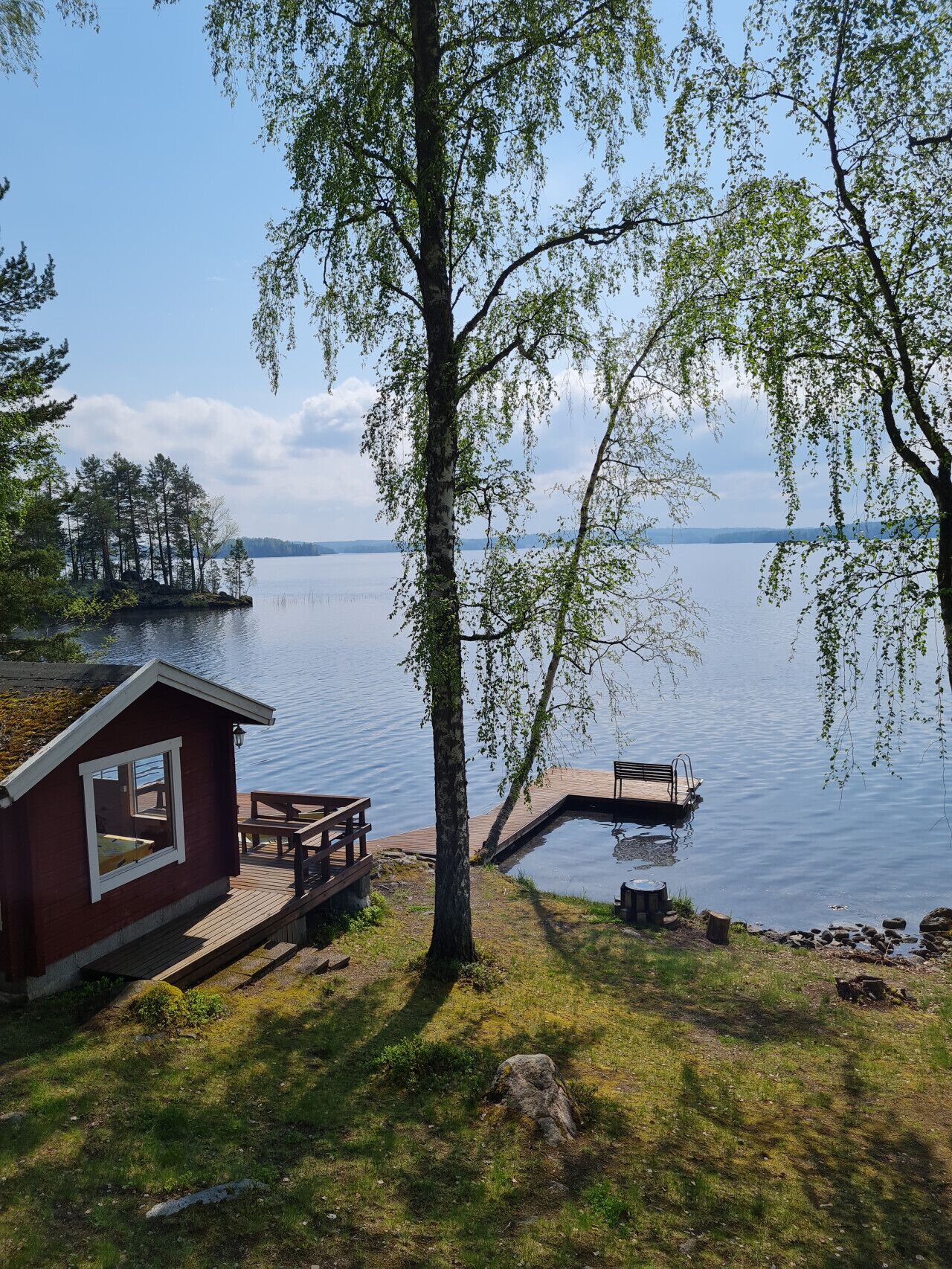 A cabin with a deck and a dock extending into a calm lake, surrounded by trees and a clear sky.
