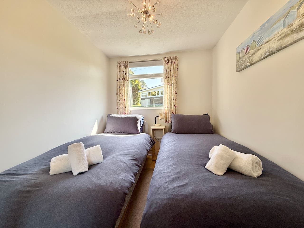 Ein kleines, gut beleuchtetes Schlafzimmer mit zwei Betten, einem Fenster mit Vorhängen, einem Bild an der Wand und einer Kristallleuchtre.