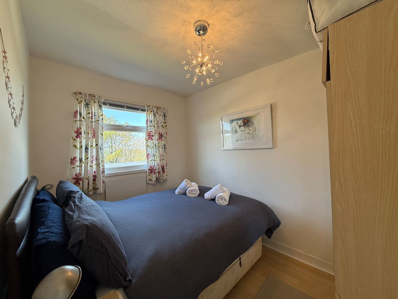 Un dormitorio con una cama, una ventana con cortinas florales, una lámpara de techo y un cuadro en la pared.