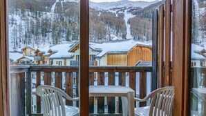 A balcony with two white plastic chairs and a small table, overlooking a snowy mountain landscape with wooden buildings. - 40m from ski slopes, 3rd floor, view ski slopes, fitness, swimming pool, sauna, balcony, tv (Les Orres)
