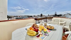 Een balkon met een ontbijtje, inclusief fruit, gebak en drankjes, met uitzicht op een stadsgezicht.