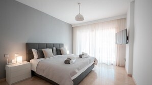 A modern bedroom with a large bed, bedside tables, a flat-screen TV mounted on the wall, and a large window with sheer curtains. - Luxury Beachfront Heated Pool & SPA & GYM–EHHouse (Loma Del Monte)
