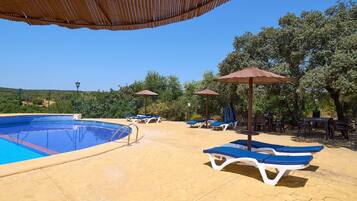 Una piscina con sillas de descanso y sombrillas, rodeada de árboles y un cielo despejado.