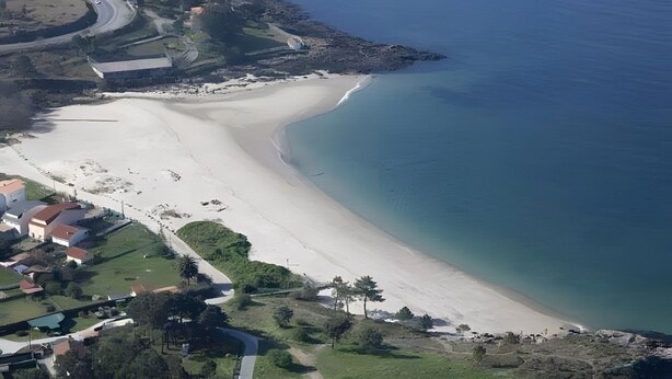 An aerial view of a coastal area with a sandy beach, a small settlement with houses, and a road curving along the coast.