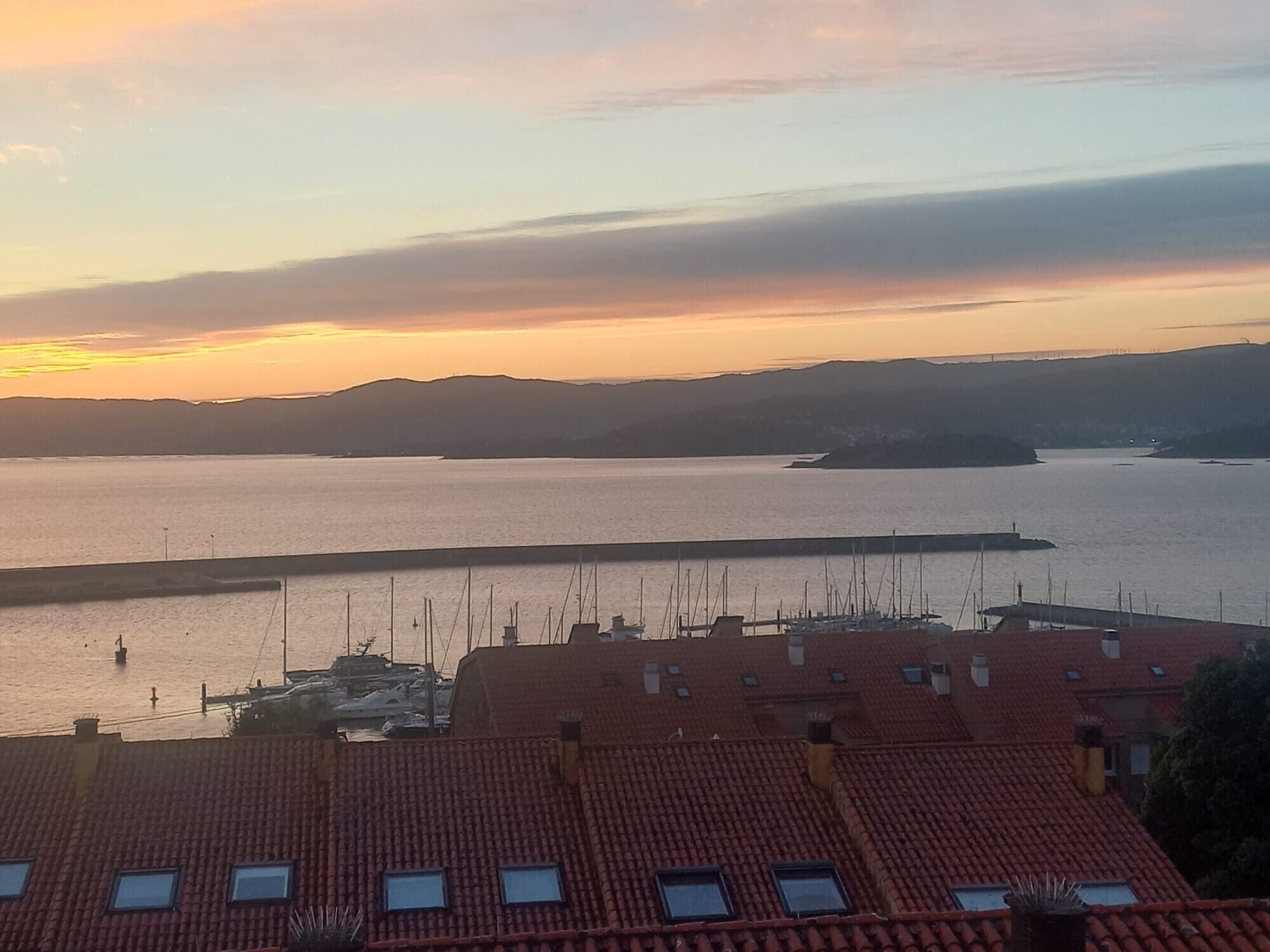 A coastal view with a marina, red-roofed buildings, and distant hills.