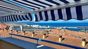 A terrace with a view of a marina and the sea, featuring striped awnings and tiled roofs.
