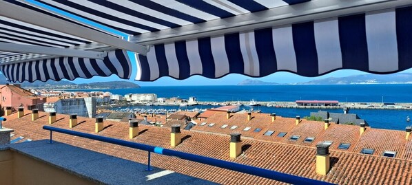 A terrace with a view of a marina and the sea, featuring striped awnings and tiled roofs.