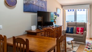 A room with a wooden dining table, chairs, a TV on a wooden stand, a sofa with nautical-themed pillows, and a view through a sliding door.