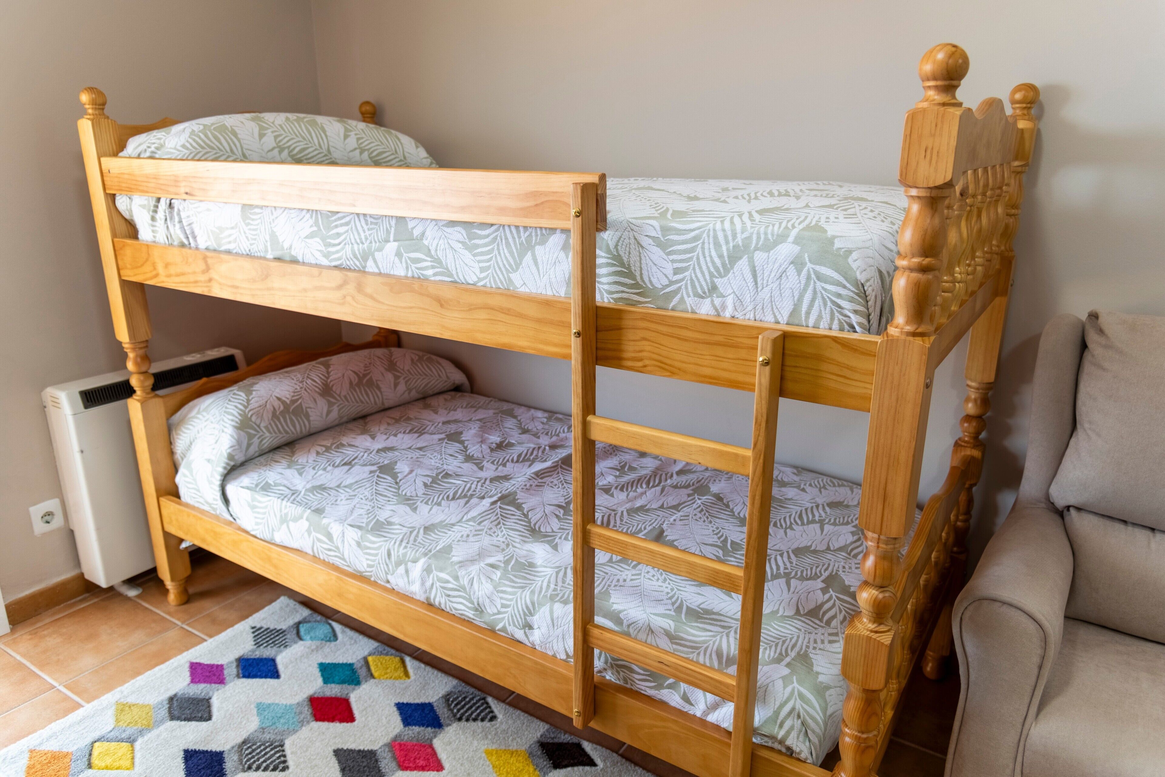 A bunk bed with a patterned bedspread, a colorful rug, and a couch in a room with tiled flooring.