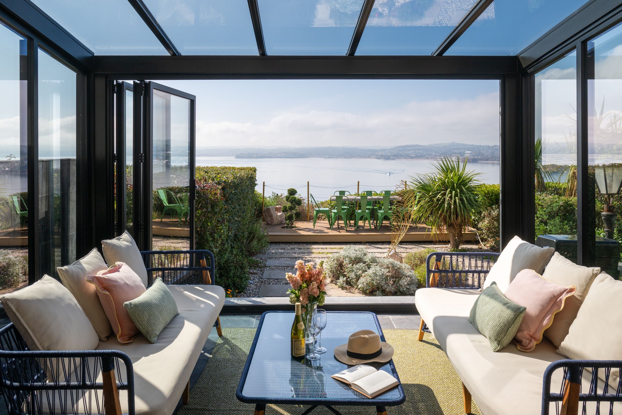 A conservatory with a glass ceiling, a sofa, a table, and a view of the sea and greenery.